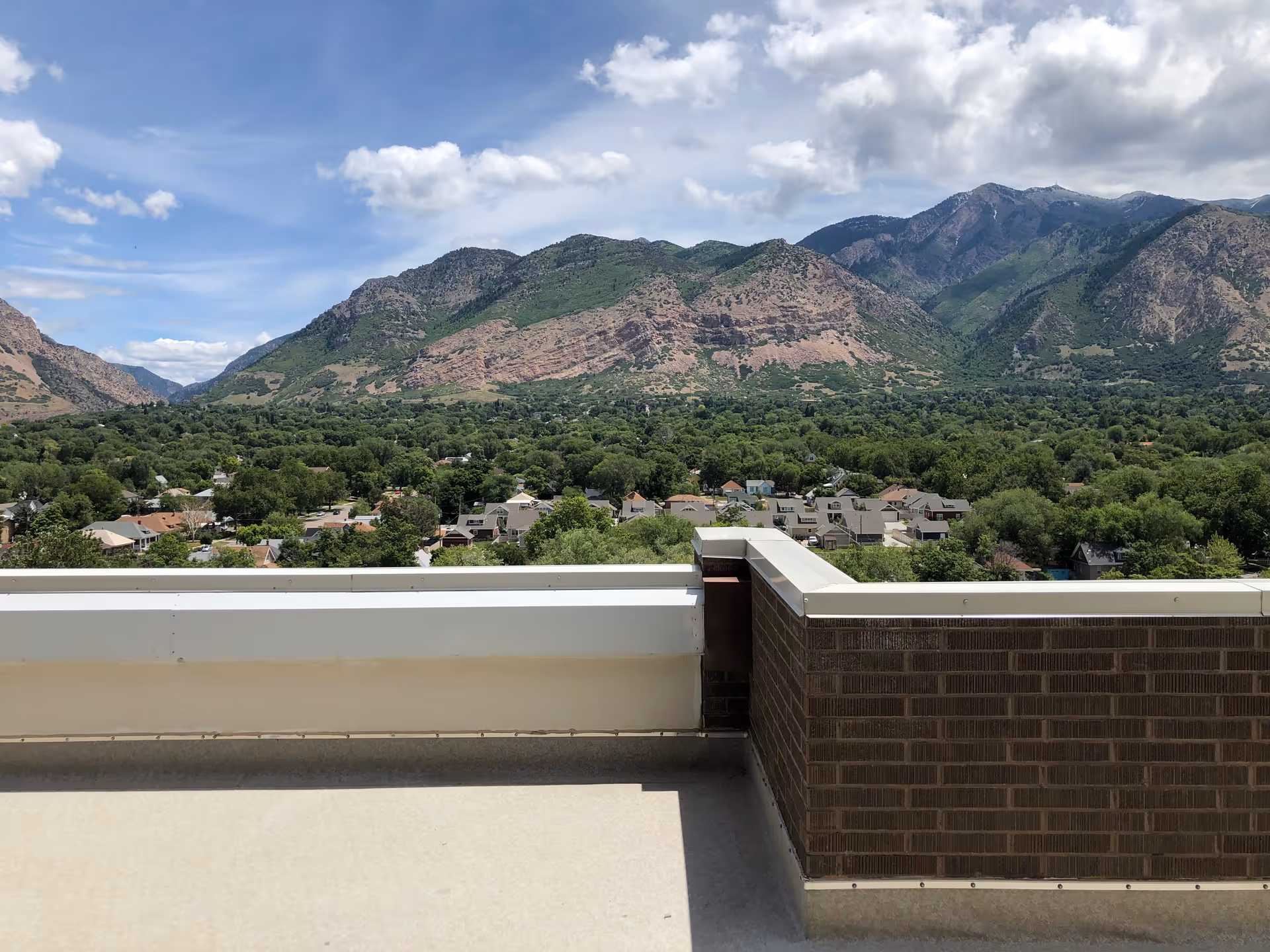 View from a rooftop terrace overlooking a residential neighborhood with many trees and houses, set against a backdrop of large, green and rocky mountains under a partly cloudy sky.
