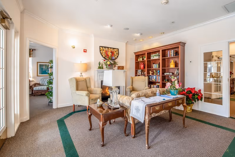 Well-lit communal sitting area with upholstered armchairs, wooden tables, a bookshelf, and floral decorations.