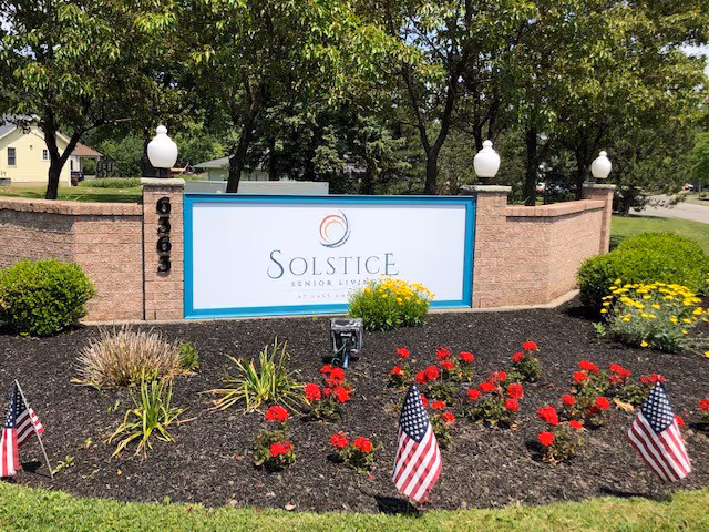 Entrance sign reading 'Solstice Senior Living' set in brick landscaping with red flowers and small American flags.