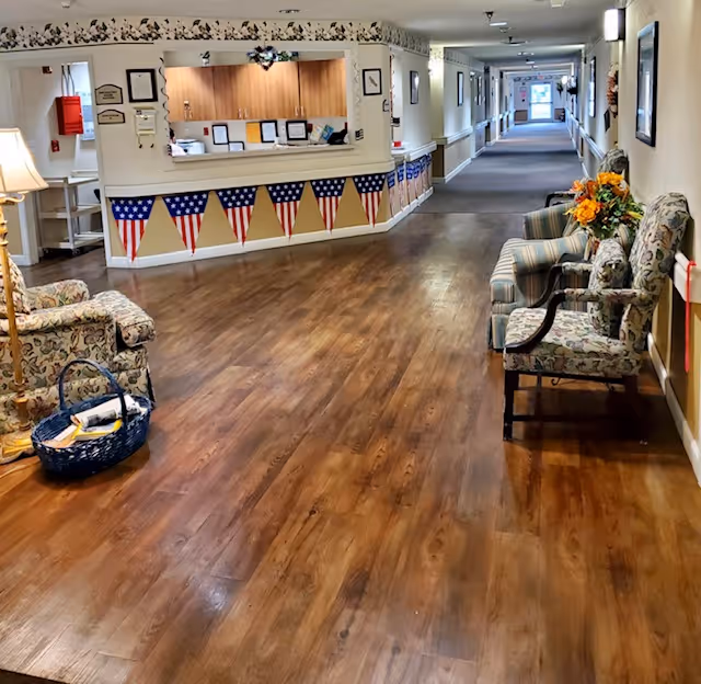 A long hallway in an assisted living facility with wood flooring and a reception desk on the left side decorated with American flag bunting. There are several upholstered chairs along the right wall, a basket with magazines on the floor near a lamp, and framed pictures on the walls. The hallway extends into the distance with doors and windows visible at the far end.
