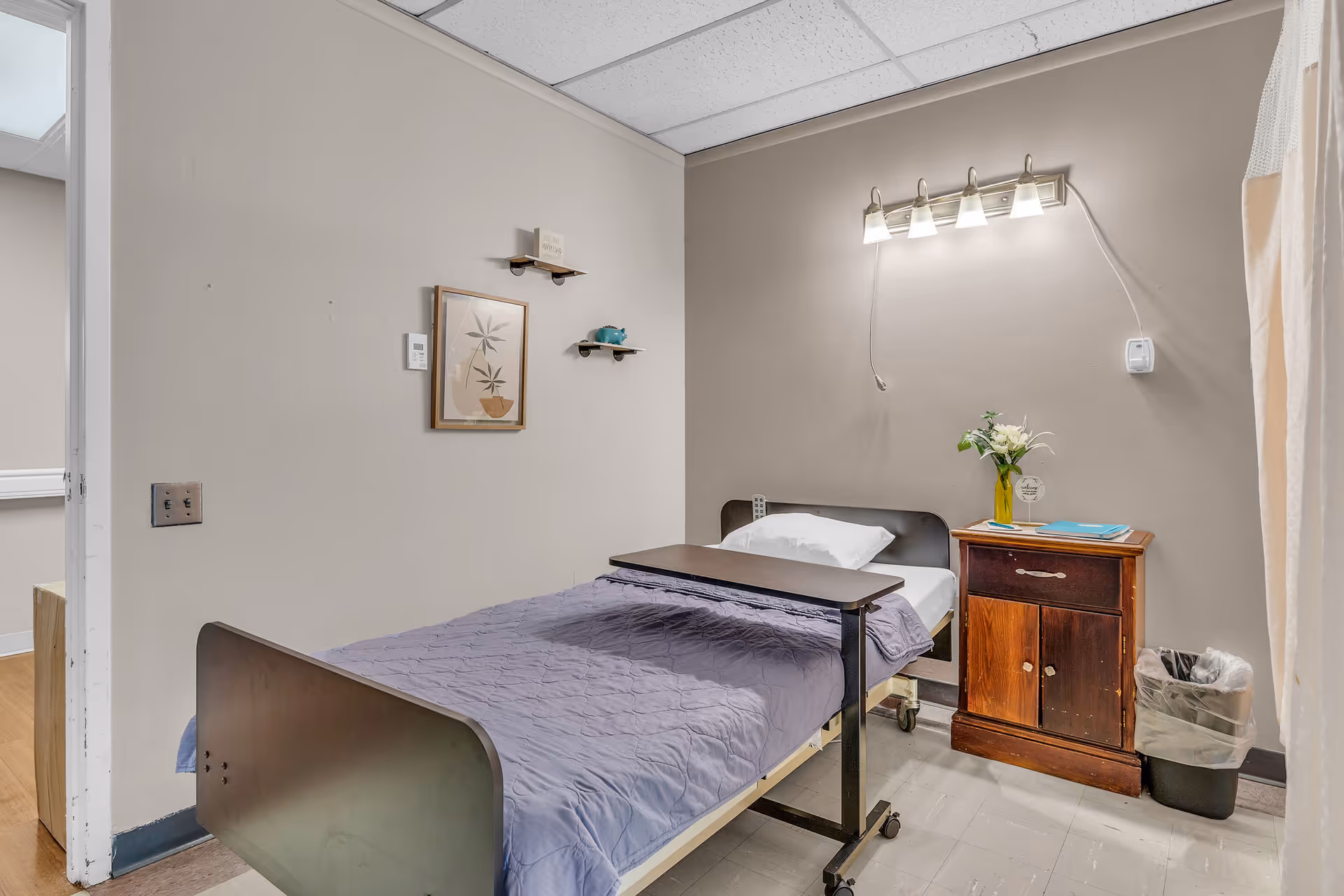 A simple patient room in a healthcare facility featuring a single bed with a purple blanket and white pillow. Above the bed is a wall-mounted light fixture with five bulbs. Next to the bed is a wooden nightstand with a vase of flowers and some documents on top. There is a small trash bin beside the nightstand. The walls are painted beige, and there is a framed picture and two small shelves on the wall opposite the bed. The ceiling has white tiles, and the floor is tiled in a light color.