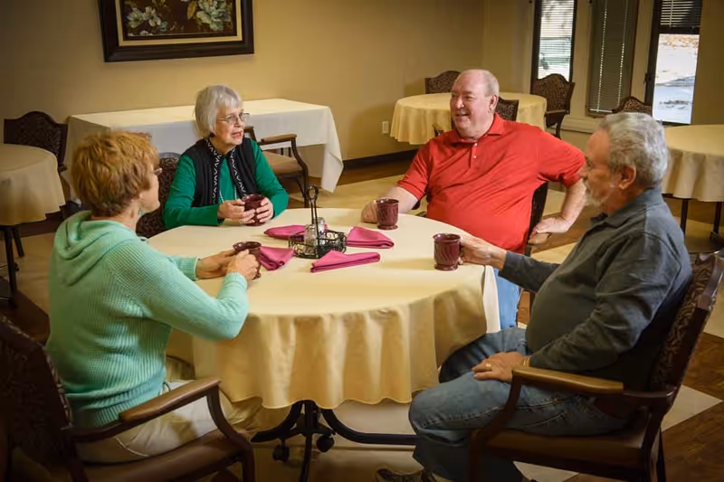 Four older adults sit around a round dining table holding mugs and chatting in a dining room.