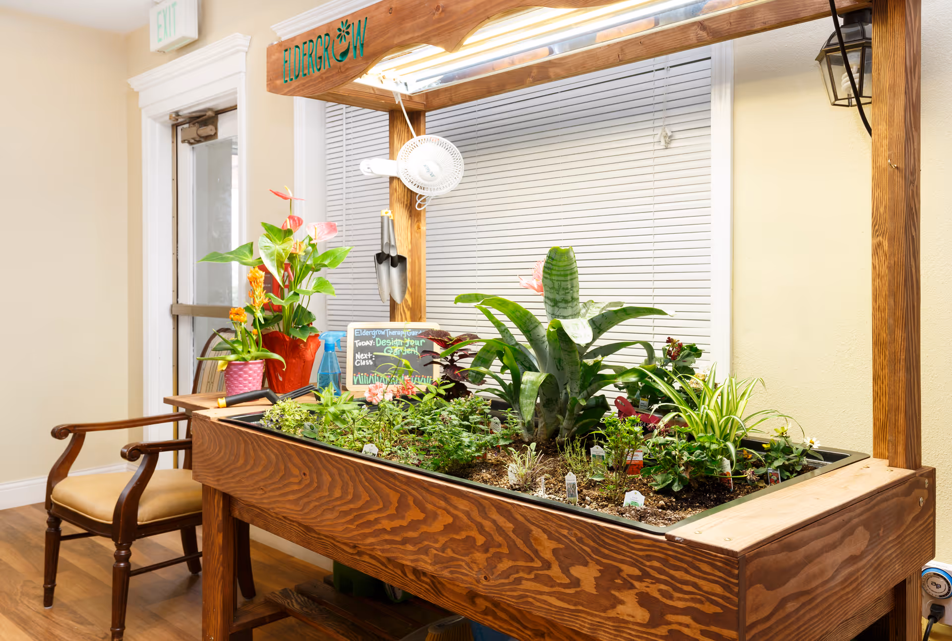 Indoor gardening table filled with various green plants and flowers, situated in a room with wooden flooring and a wooden chair nearby. A wooden frame above the table has the word 'Eldergrow' on it, and gardening tools hang from the frame. A small chalkboard sign with gardening notes is placed among the plants. A window with closed blinds is in the background.