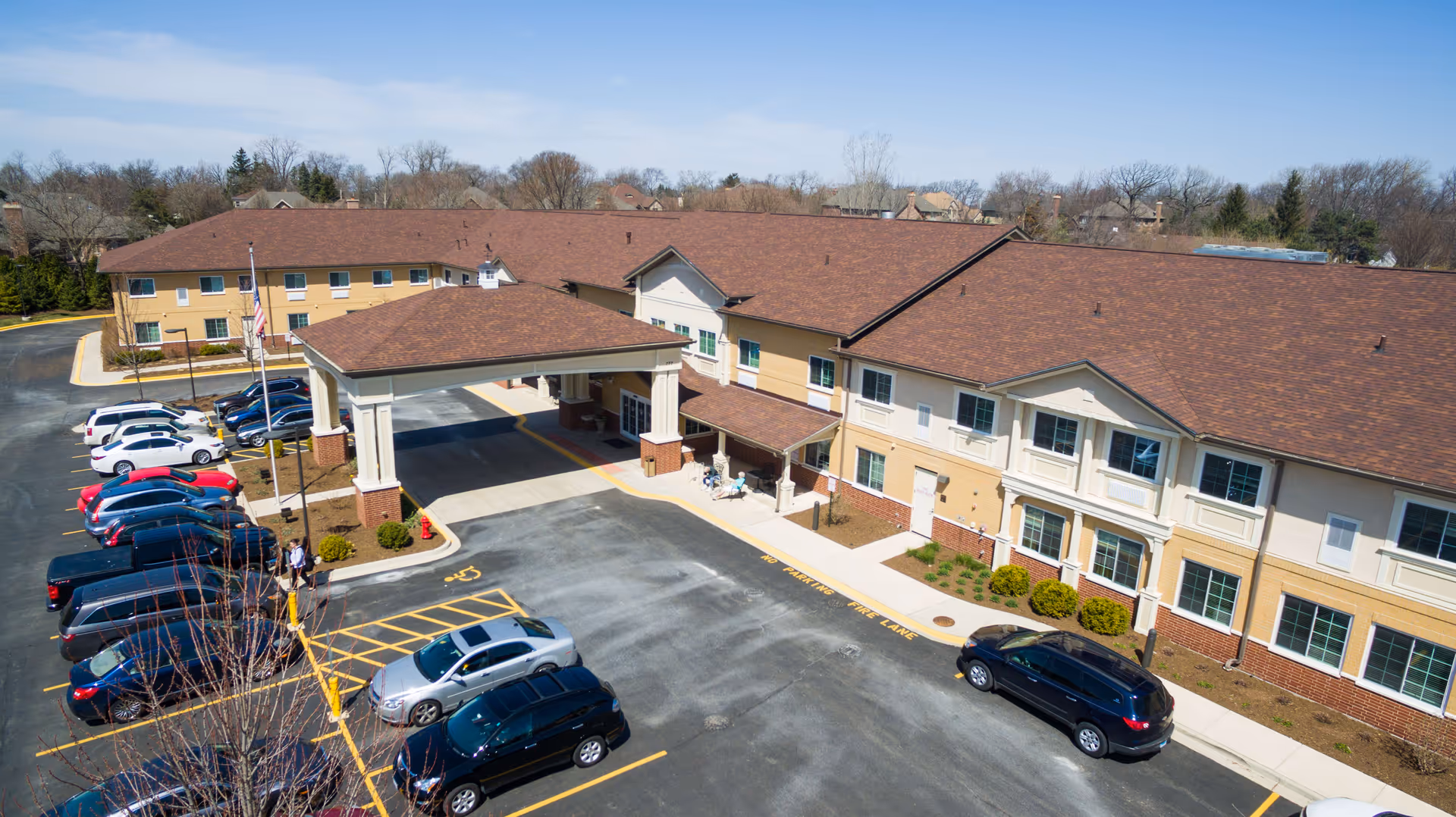 Aerial view of Cedar Lake Assisted Living and Memory Care building with a covered entrance and parking lot filled with cars. The building is two stories with a brown roof and beige exterior walls. Trees and houses are visible in the background under a clear sky.