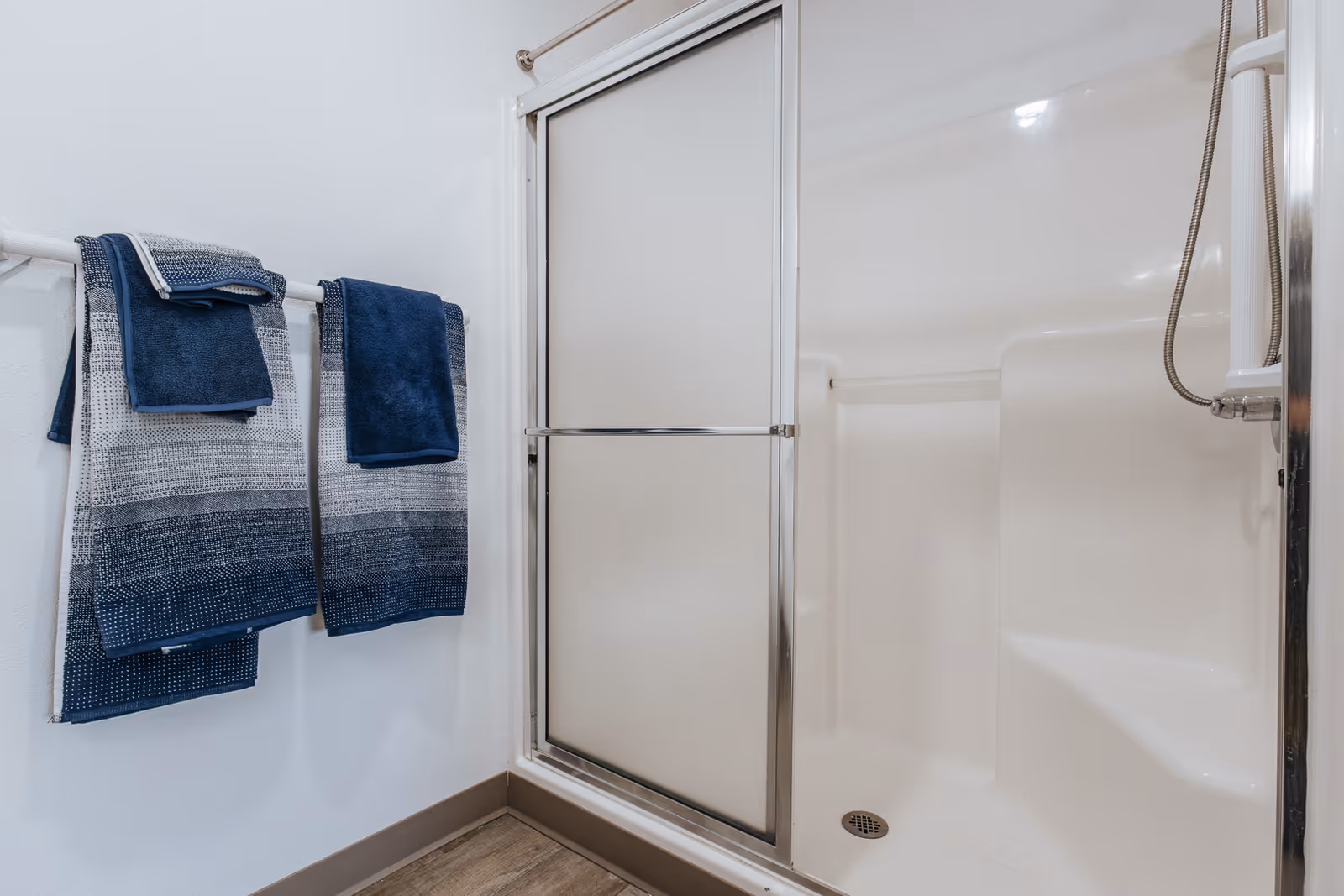 A clean bathroom shower area with a sliding glass door and built-in seating. Two towel racks on the adjacent wall hold neatly folded blue and gray towels. The floor has a wood-like finish.