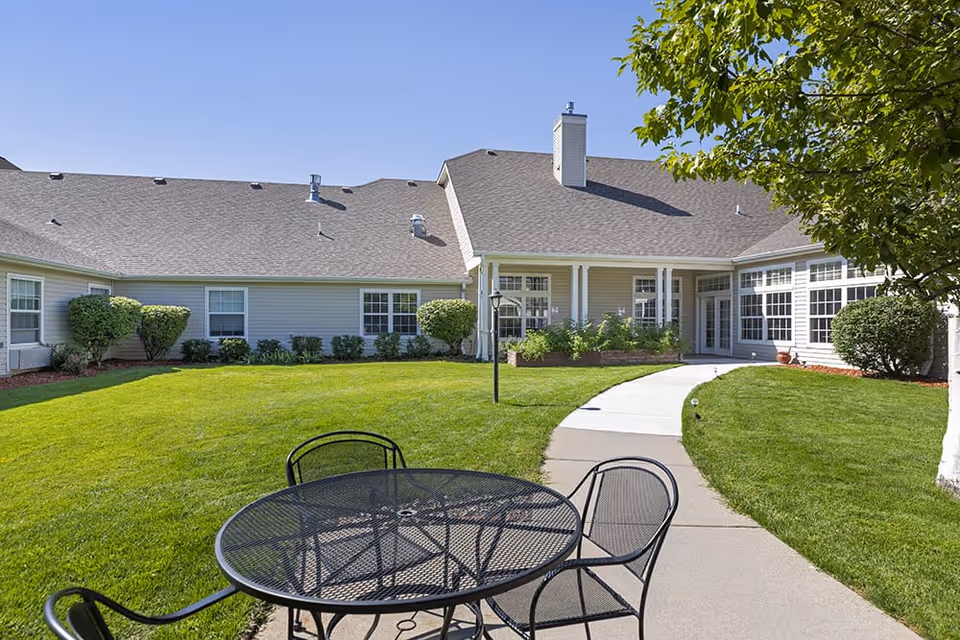 Sunlit courtyard with a metal patio table and chairs, a curved concrete path, manicured lawn, and a single-story building with large windows.