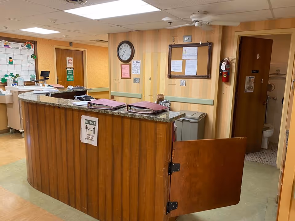 Wooden reception desk/nurse station in a senior care facility hallway with a clock, bulletin board, paperwork, and an open restroom door.