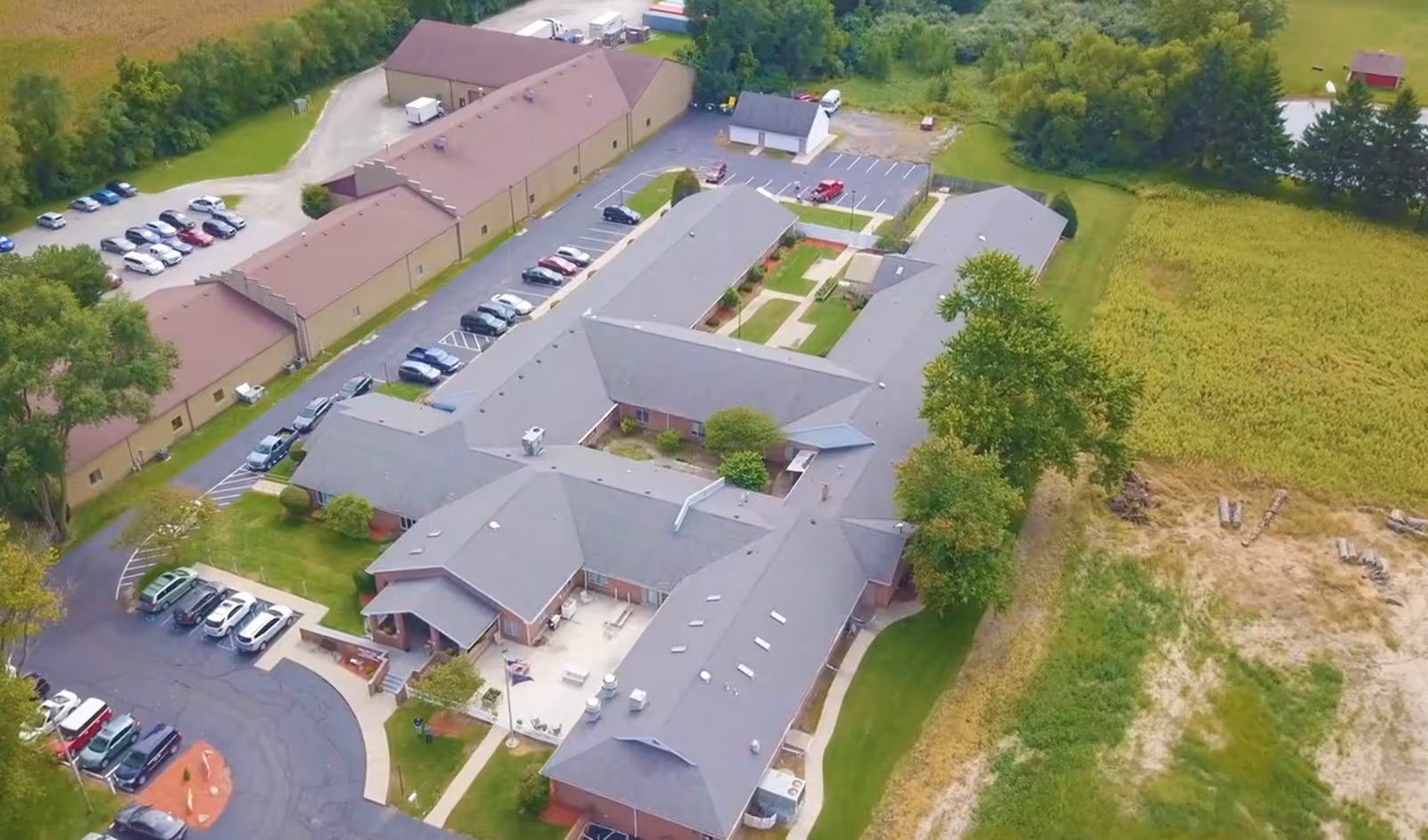 Aerial view of Swanton Valley Rehabilitation and Healthcare Center showing multiple connected buildings with gray roofs, surrounding parking lots with cars, green lawns, trees, and adjacent fields.