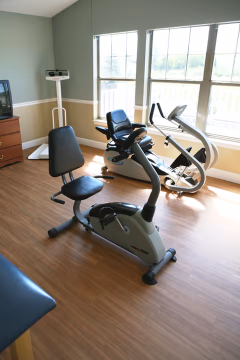 A small exercise room with two stationary exercise bikes positioned near large windows letting in natural light. A scale and a wooden dresser with a TV on top are visible in the corner. The room has wood flooring and walls painted in two tones, green above and beige below with white trim.