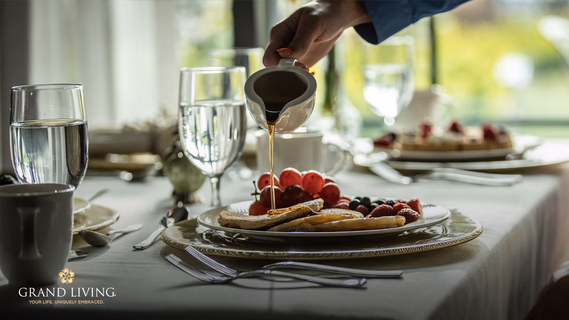 A close-up view of a dining table set with plates of pancakes topped with fresh berries. A hand is pouring syrup onto the pancakes. The table is elegantly set with glasses of water, coffee cups, and silverware, with a softly lit background.
