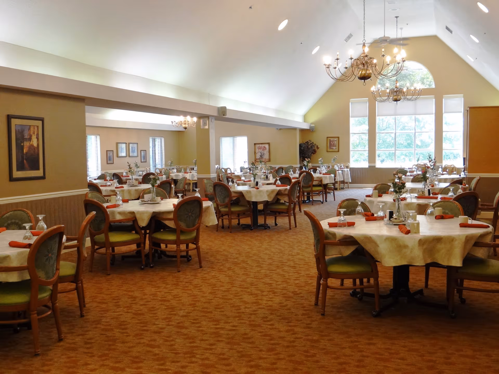 A spacious dining room with multiple round tables covered with beige tablecloths, each set with glassware, napkins, and small floral centerpieces. The room has green cushioned wooden chairs, large windows letting in natural light, chandeliers hanging from a vaulted ceiling, and framed artwork on the walls.