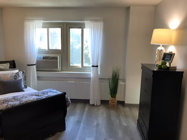 A bedroom with a black wooden bed frame and patterned bedding, a window with white curtains and an air conditioning unit, a tall black dresser with a lamp and decorative items on top, and a potted plant on the floor near the window.