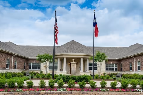 Front view of a single-story brick senior living building with a columned entrance, American and Texas flags, and landscaped gardens in front.