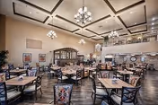 Spacious dining room in a senior living facility with multiple wooden tables and chairs arranged neatly. The room features high ceilings with decorative beams, elegant chandeliers, framed artwork on the walls, and a balcony overlooking the area. The floor is wooden, and there are plants placed around the room for decoration.