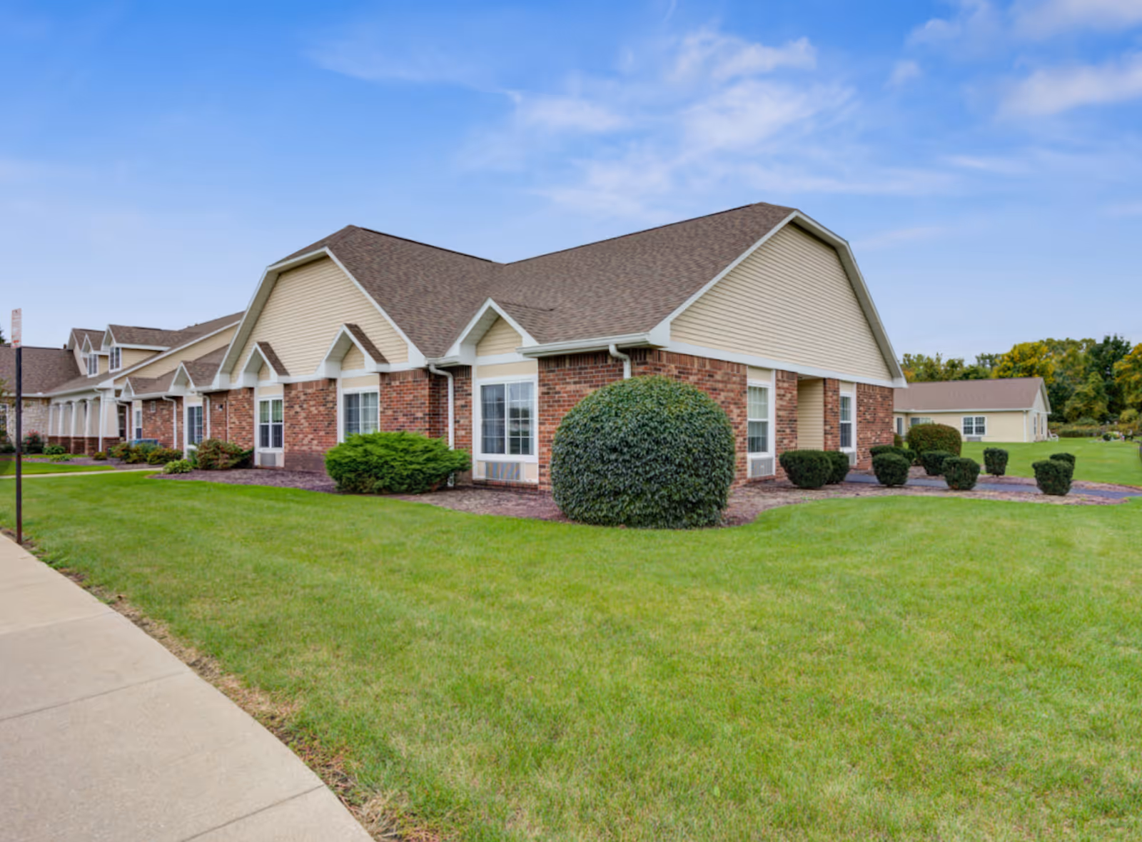 Exterior view of a single-story senior living community building with brick and beige siding, surrounded by well-maintained green lawns and shrubs under a partly cloudy blue sky.