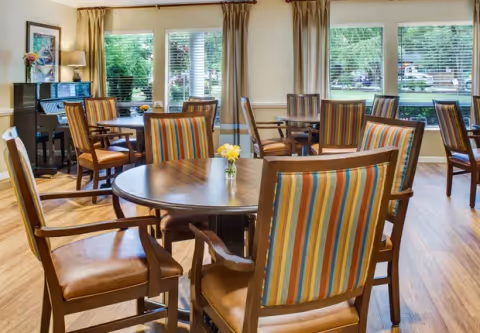 Bright memory-care dining room with round wooden tables, striped upholstered chairs, large windows, and a piano in the corner.