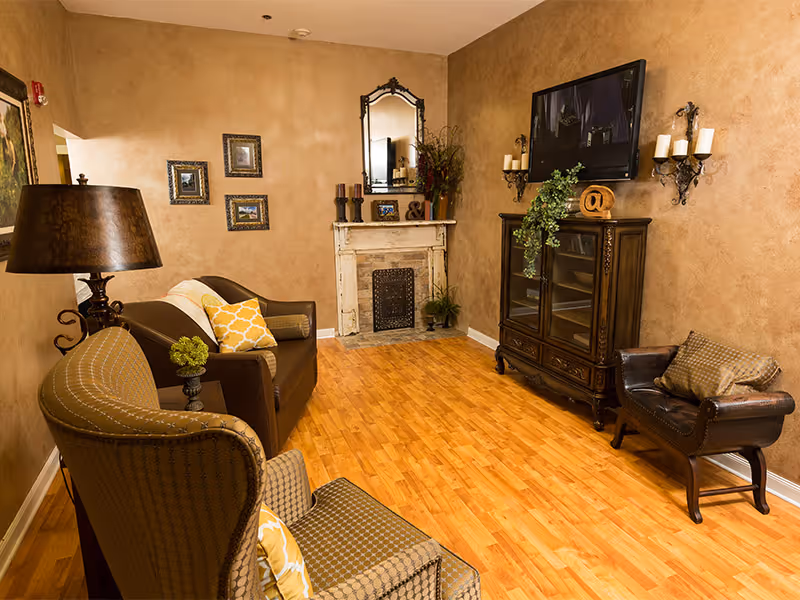 Cozy living room with brown upholstered chairs and sofa, a decorative fireplace, wooden cabinet with a wall-mounted TV, and warm-toned walls and hardwood floor.