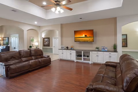 A spacious living room with two brown leather sofas facing a wall-mounted flat screen TV. The room has wooden flooring, a ceiling fan with lights, and white cabinetry under the TV. There are decorative plants and framed pictures on the cabinets and walls. The room has arched doorways leading to other areas of the facility.