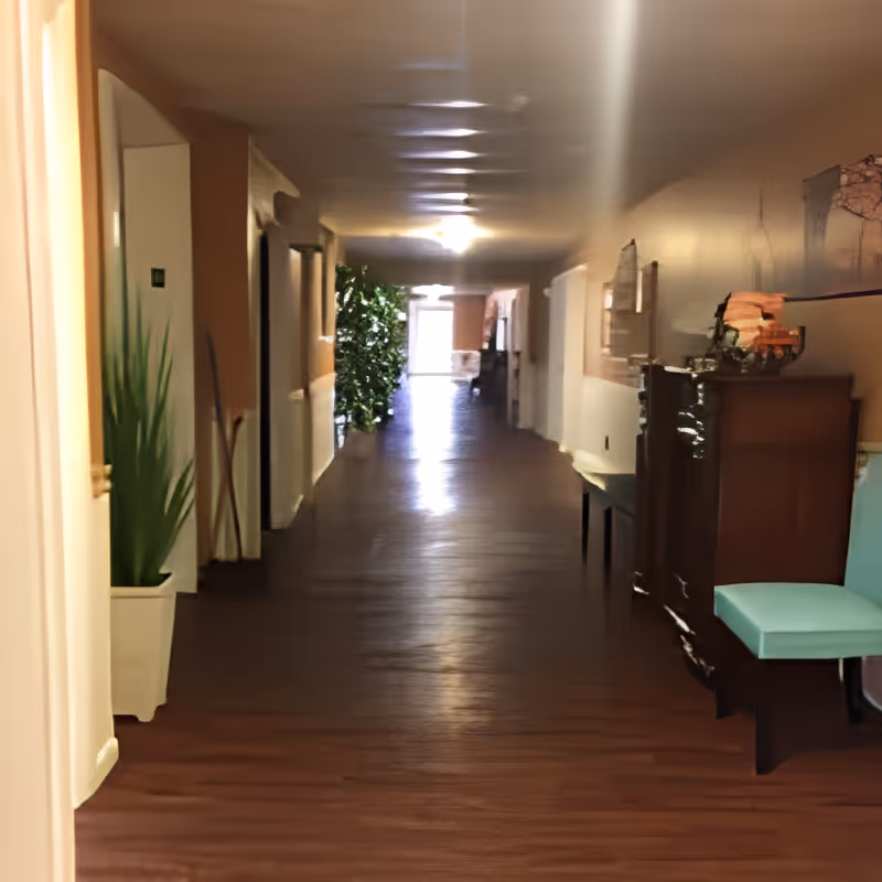 A long indoor hallway with wooden flooring and beige walls, featuring a potted plant on the left and a wooden cabinet with a light blue cushioned chair on the right. The hallway is lit by ceiling lights and has doors and decorations along the walls, leading to a bright exit at the far end.