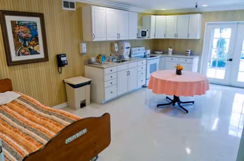 Interior of a senior living room with a bed, kitchenette, and round pink table.