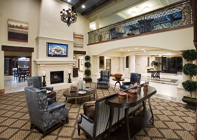 Spacious and elegant senior living facility common area with patterned carpet, comfortable upholstered chairs and sofas arranged around a central coffee table, a large fireplace with a mounted TV above it, decorative plants, and a wrought iron railing on the upper level balcony.