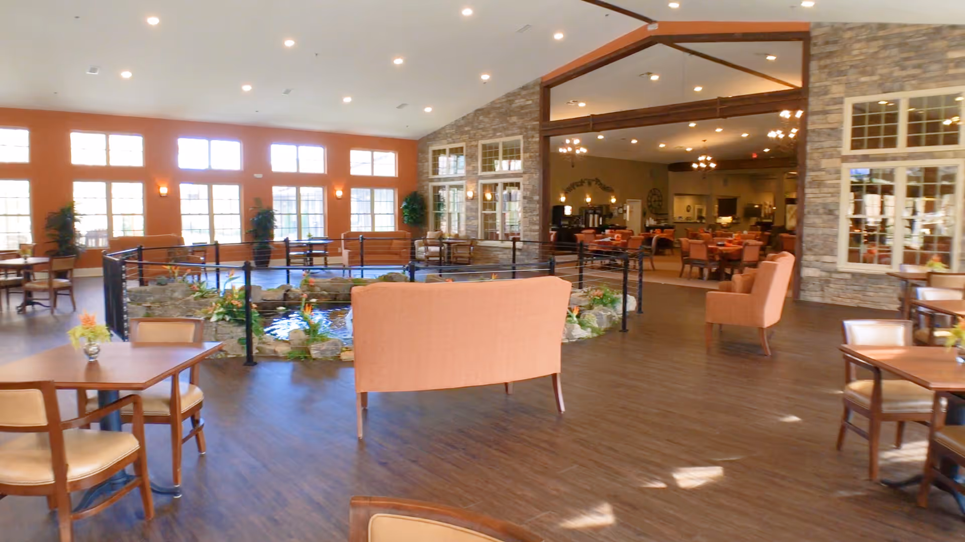 Spacious and well-lit common area in a senior living facility with large windows, wooden flooring, multiple seating arrangements including chairs and sofas, and a central indoor water feature surrounded by plants and rocks. The area opens into a dining room with tables and chairs, and the walls feature stone accents and warm lighting.