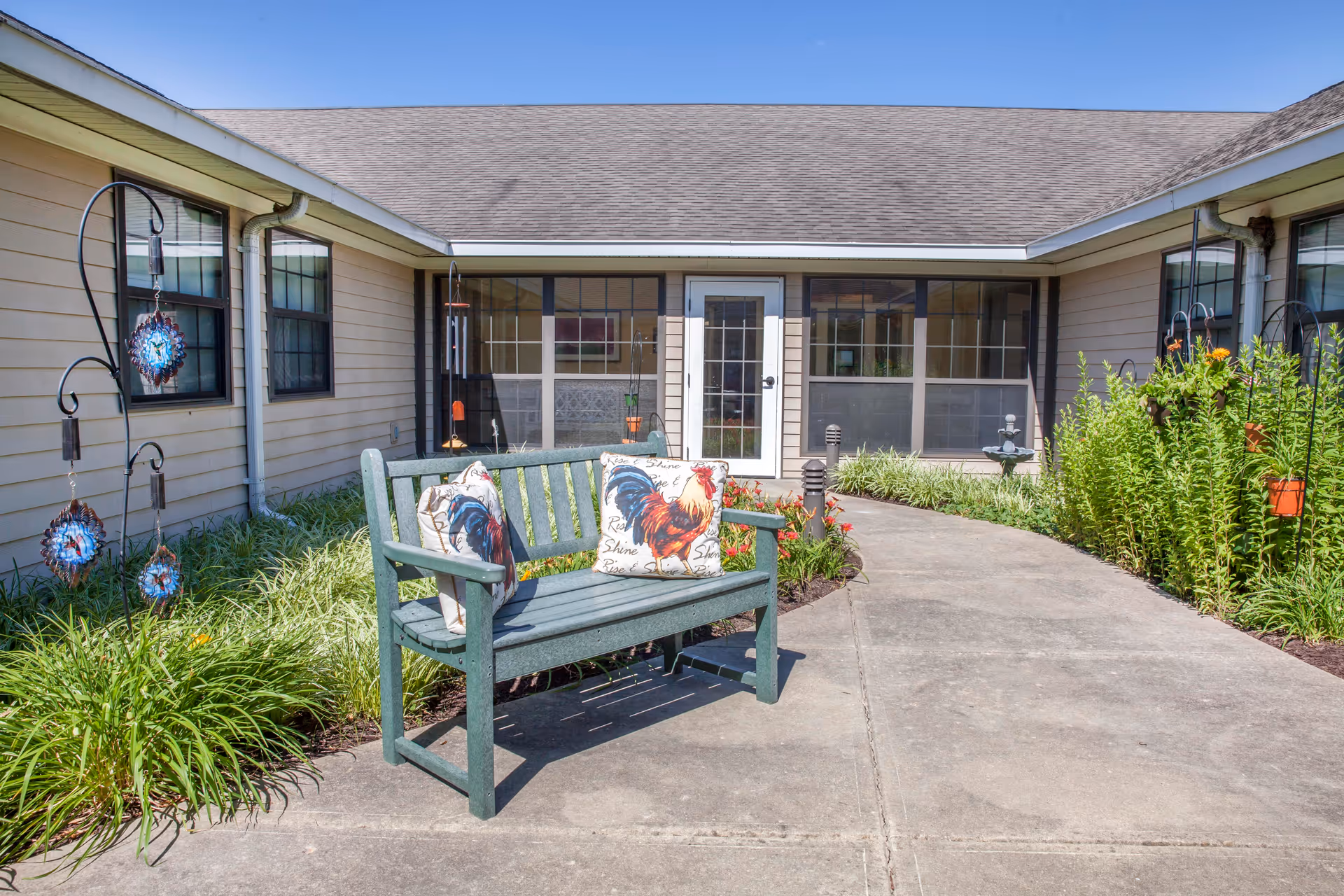 Outdoor courtyard with a green bench and rooster-printed cushions on a paved walkway leading to a screened entrance of the building.