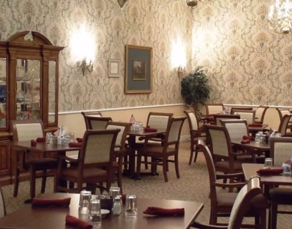 A dining room with multiple wooden tables and chairs arranged neatly. Each table has glasses, napkins, and small condiment containers. The walls are decorated with patterned wallpaper, framed pictures, and wall sconces providing soft lighting. There is a wooden cabinet with glass doors displaying items on the left side and a potted plant in the corner.