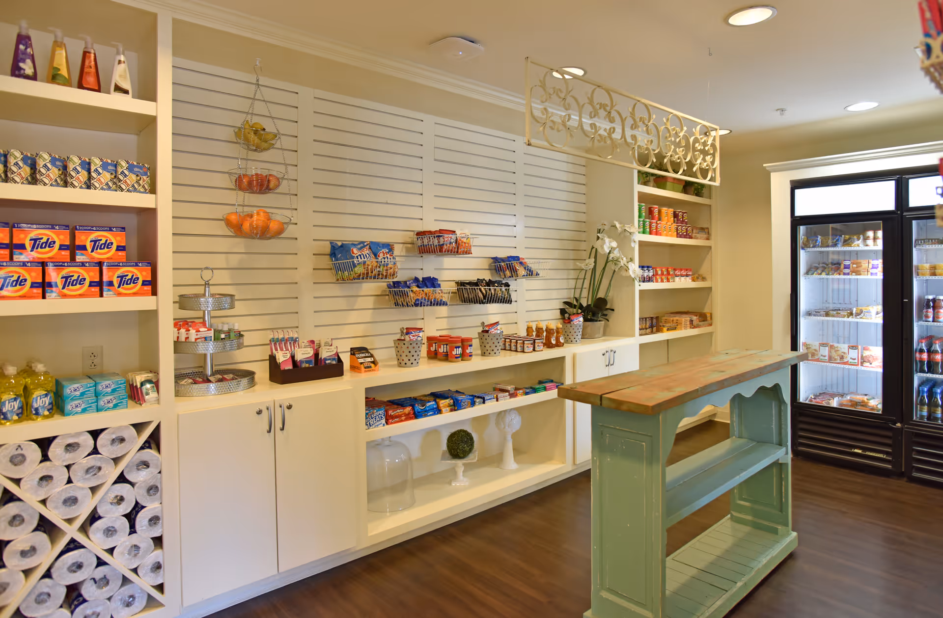 Interior view of a small convenience or pantry area with shelves stocked with household items like paper towels, laundry detergent, dish soap, snacks, and canned goods. There is a green wooden island in the center and a refrigerator with glass doors containing beverages and food items. The space has warm lighting and wooden flooring.