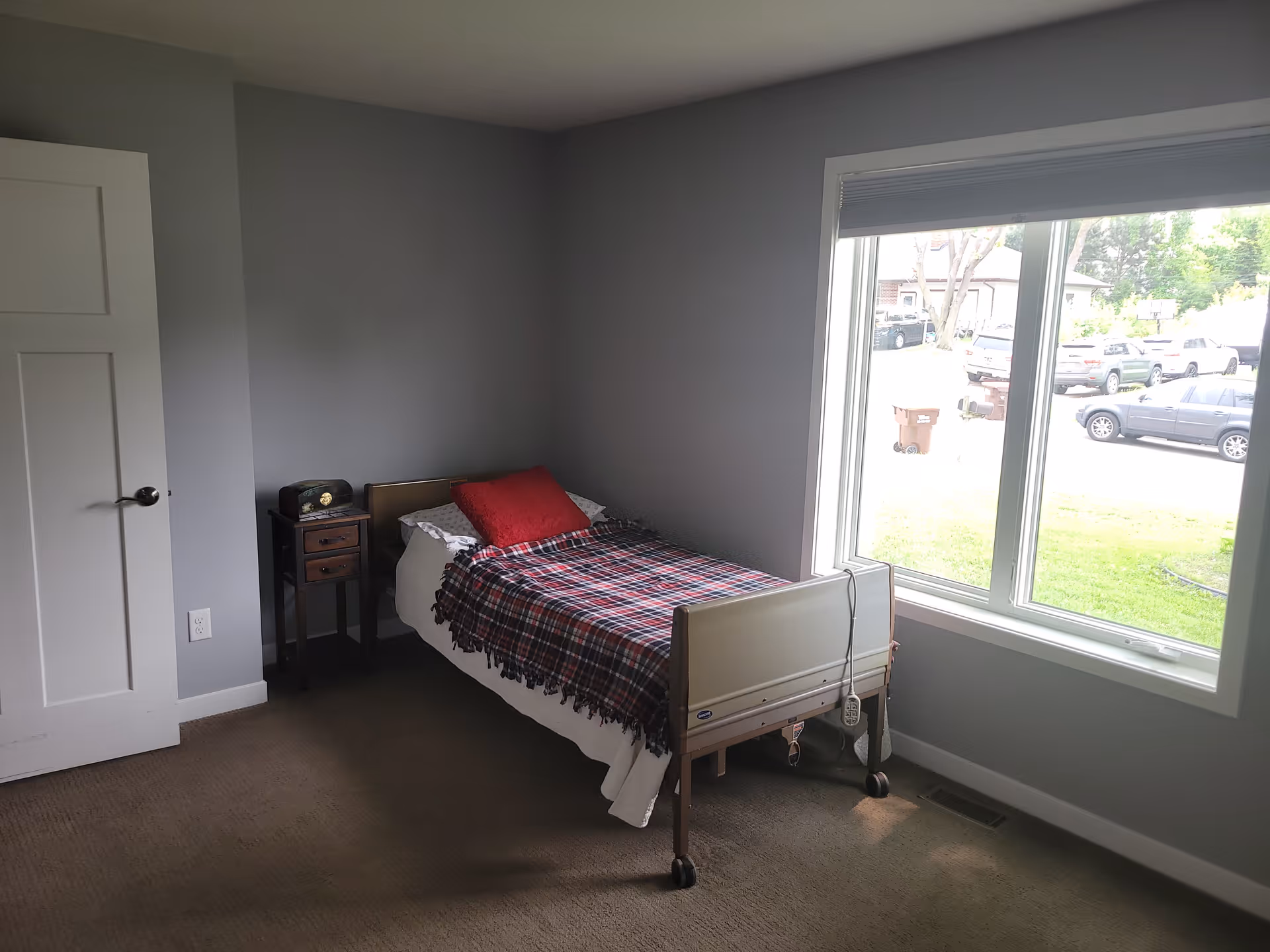 A simple single hospital-style bed with a red pillow and plaid blanket beside a window and small bedside table in a gray-painted bedroom.