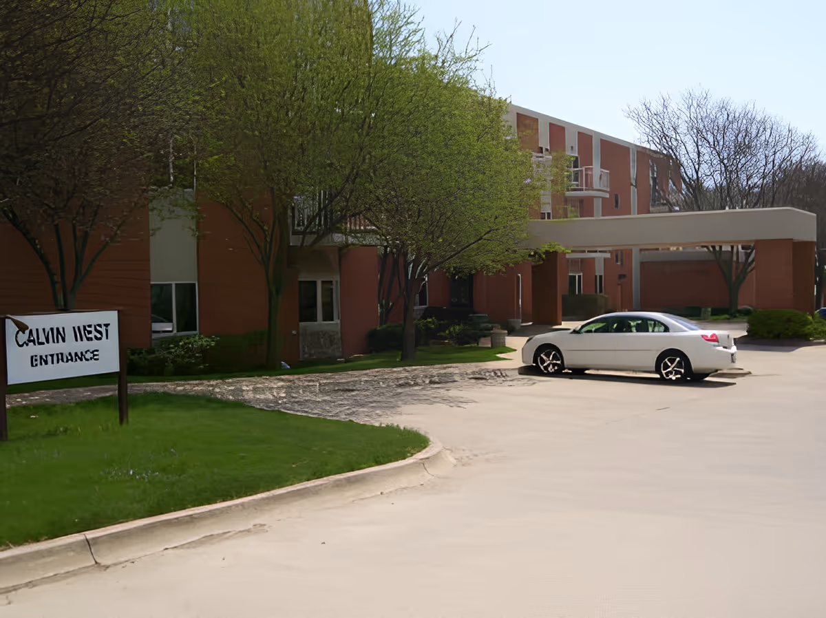 Exterior view of a senior living facility named Calvin Community showing a red brick building with multiple floors, a covered entrance, a white car parked in front, green trees, and a sign that reads 'Calvin West Entrance'.