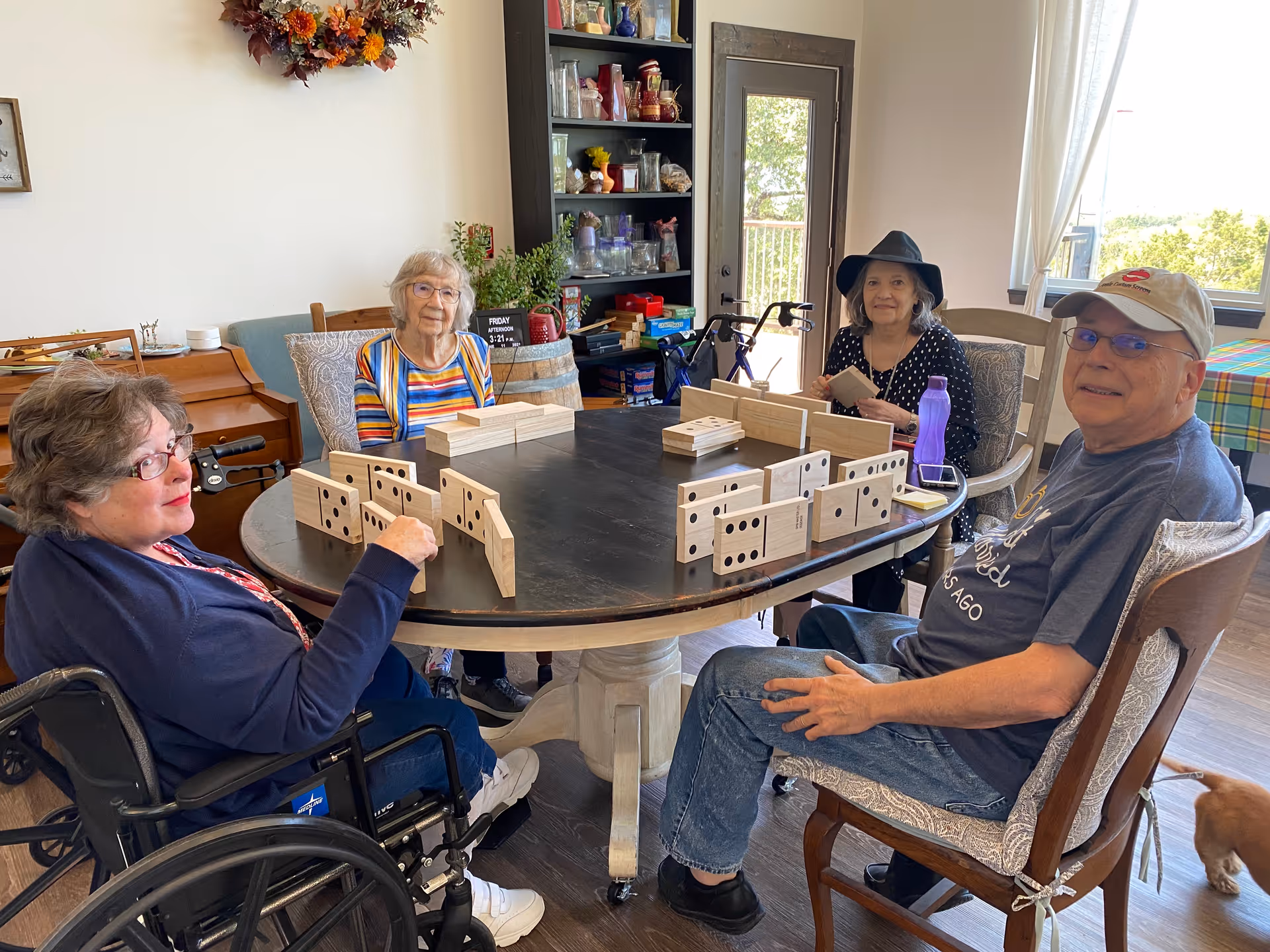 Four elderly individuals sitting around a round table playing a game with large wooden dominoes in a well-lit room. One person is in a wheelchair, and there is a shelf with various items and a door leading outside in the background. A small dog is partially visible near the bottom right corner.