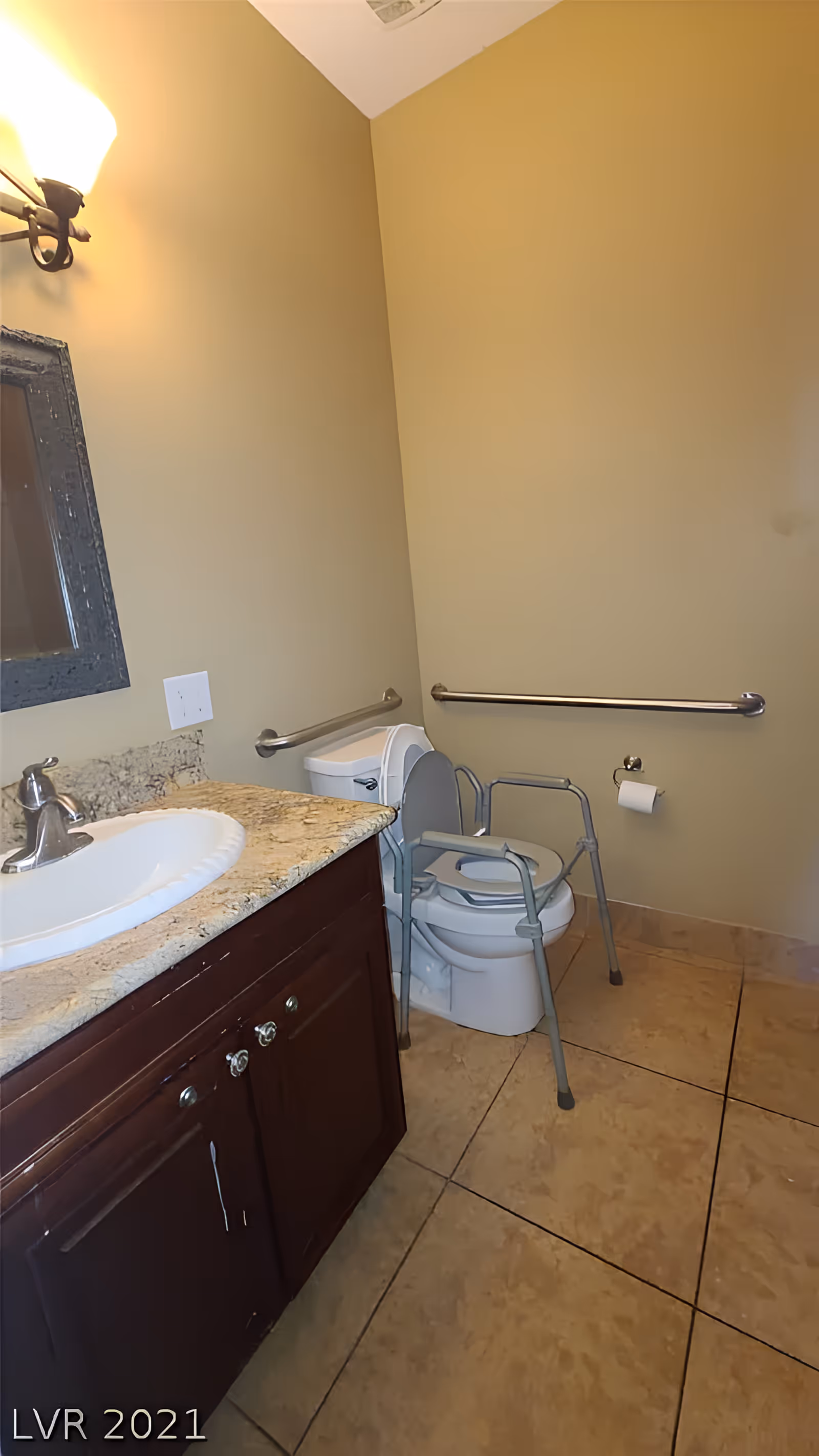 A bathroom with beige walls and tiled floor. There is a wooden vanity with a granite countertop and a white sink on the left side. Above the sink is a mirror and a wall-mounted light fixture. On the right side, there is a toilet with a raised toilet seat and arm supports, along with grab bars on the walls for accessibility. A toilet paper holder is mounted on the wall next to the toilet.