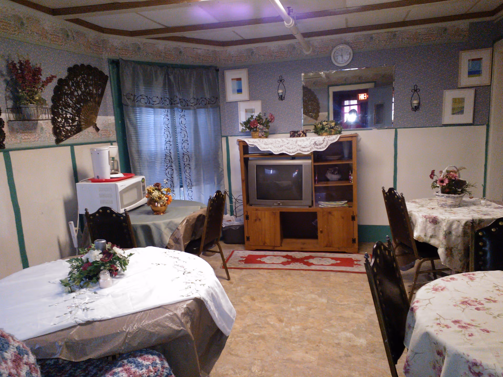 A cozy common room with multiple round tables covered with floral and plain tablecloths, each decorated with flower arrangements. There is a wooden cabinet with an old-style TV and a lace runner on top. A microwave and coffee maker are placed on a small table near a window with sheer curtains. The walls have framed artwork, a decorative fan, and sconces, with a clock above the TV. The floor is covered with a light-colored linoleum.