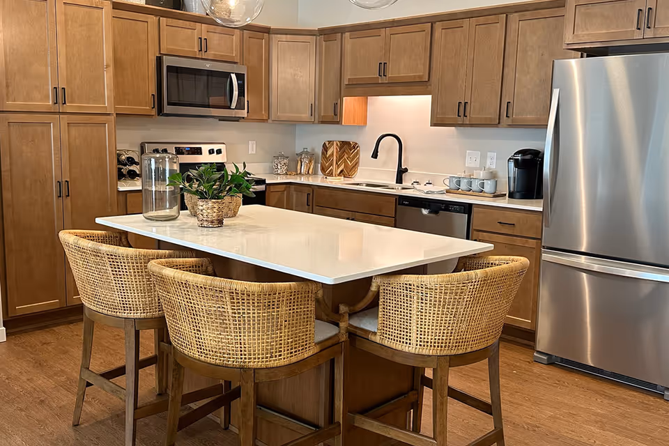 Modern kitchen with wooden cabinets, stainless steel refrigerator, microwave, and dishwasher. A white island countertop with four wicker chairs surrounds it. There are decorative plants and jars on the countertop.