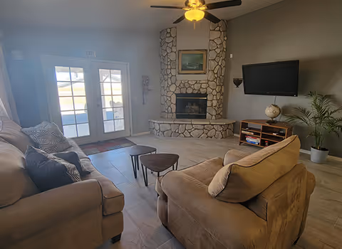 A cozy living room with two beige sofas facing a stone fireplace. Above the fireplace is a framed painting. To the right of the fireplace is a mounted flat-screen TV and a wooden media console with a globe and some books. A potted plant is placed in the corner. Double glass doors with an exit sign above them lead outside, and a ceiling fan with a light fixture is on the ceiling.