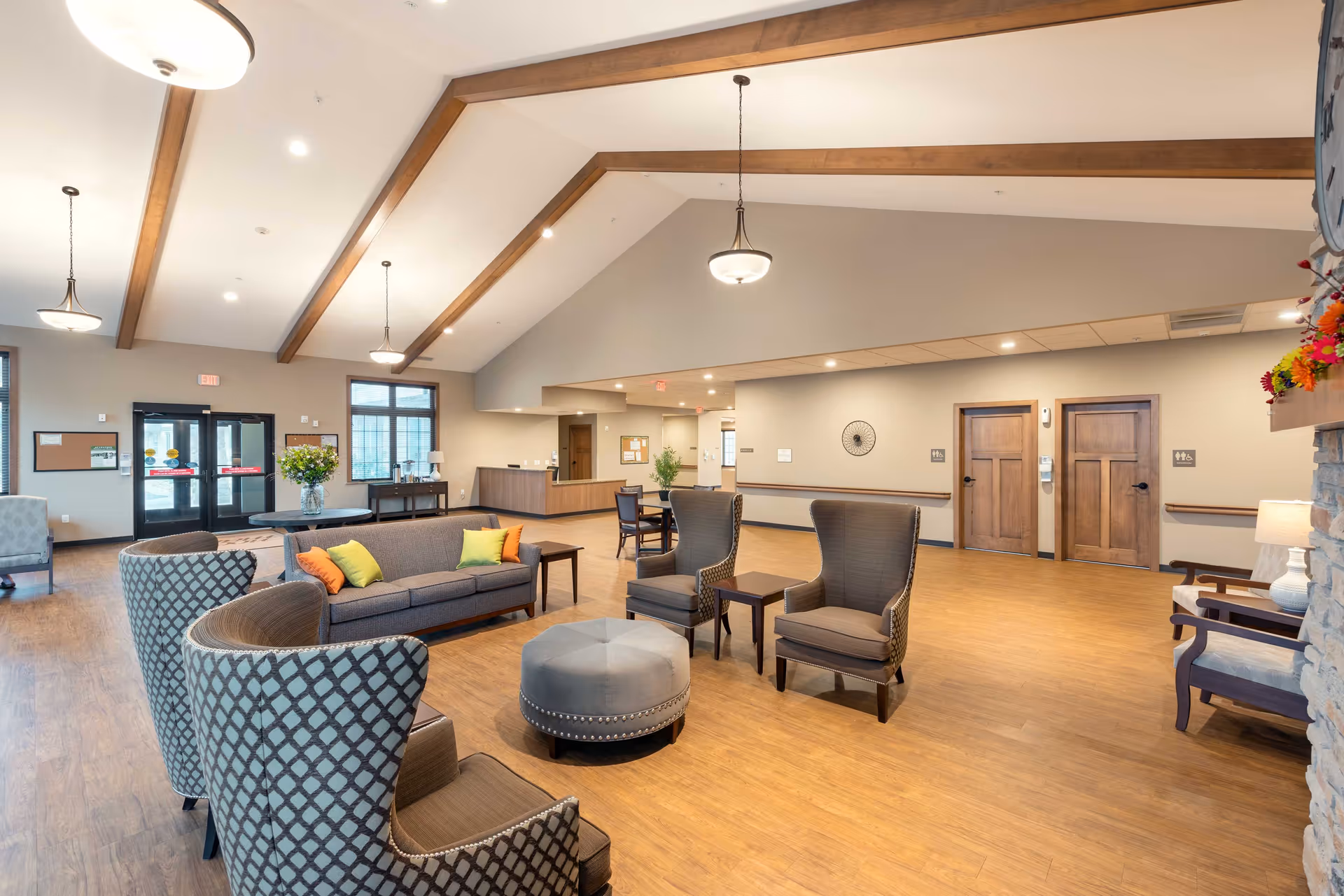 Spacious furnished common lounge in a senior living facility with sofas and armchairs, vaulted ceiling with wooden beams, and a reception desk in the background.