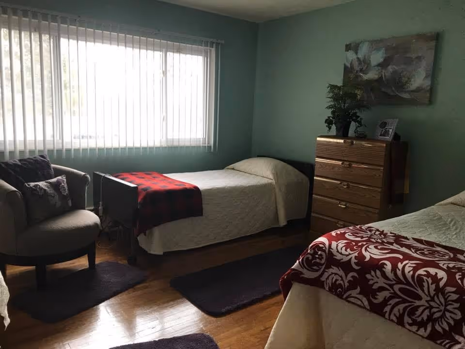 A cozy bedroom with two single beds covered in white quilts and decorative blankets, a wooden chest of drawers with a plant and picture frame on top, a comfortable armchair with a pillow, and a large window with vertical blinds letting in natural light.