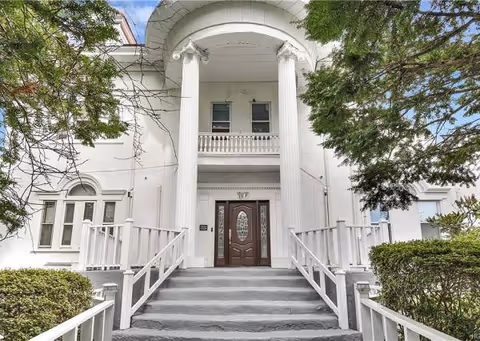 White building entrance with tall columns, stairs, and a decorative door flanked by trees.