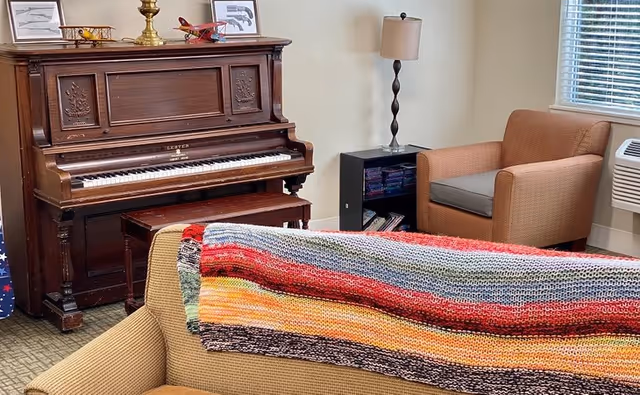 A cozy living room area with an upright piano, armchairs, a sofa draped with a colorful knitted blanket, and a lamp by a window.
