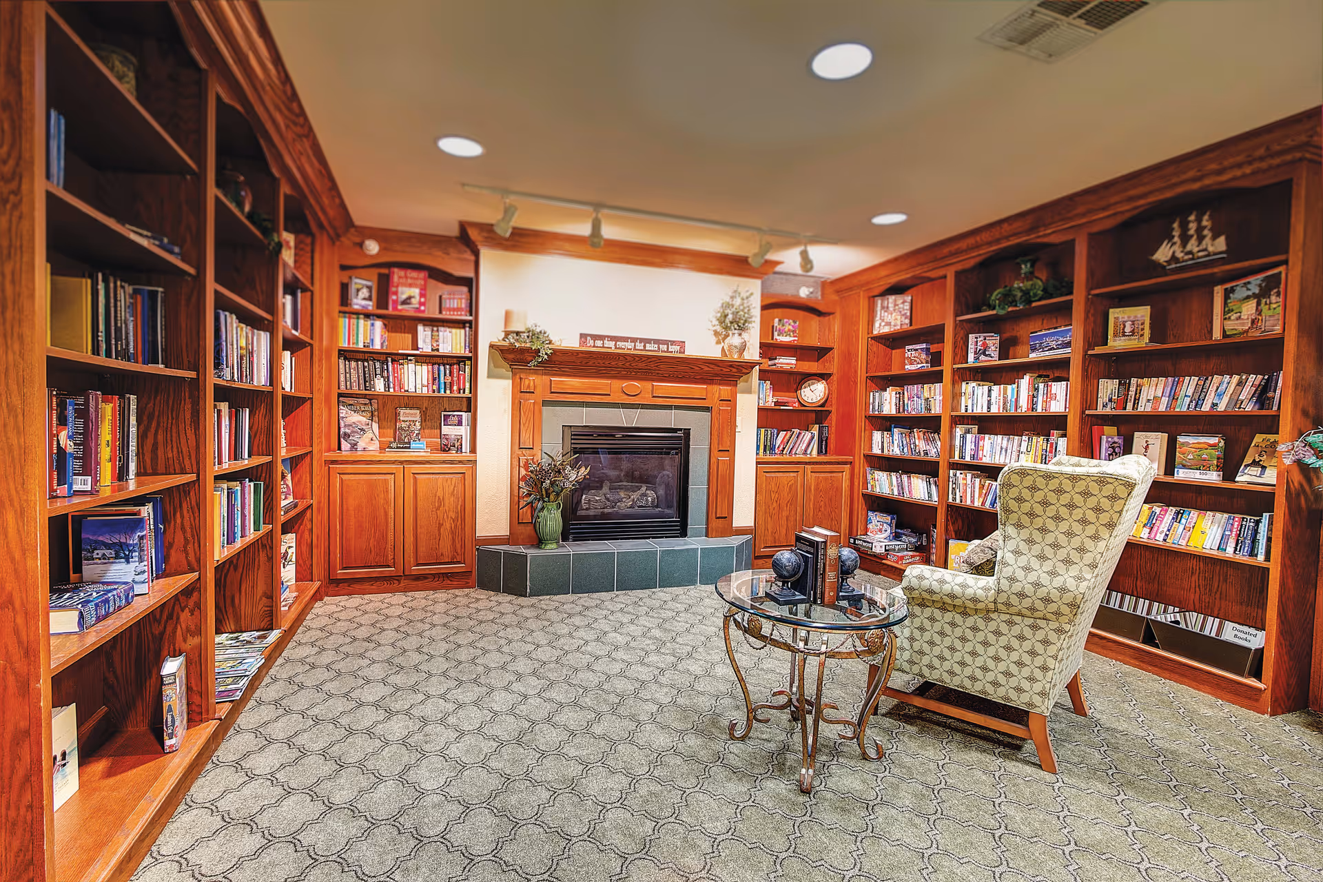 A cozy library room with wooden bookshelves filled with books surrounding a fireplace. There is a patterned armchair and a glass-top round table with decorative items on it. The room has a carpeted floor and warm lighting.