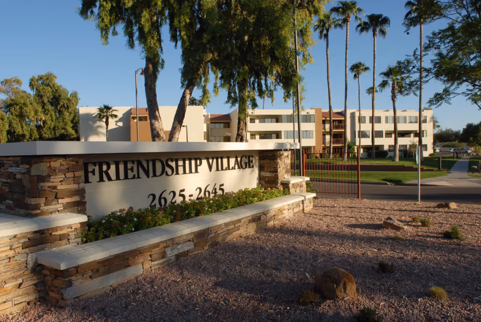 Stone entrance sign reading 'FRIENDSHIP VILLAGE' in front of a multi-story senior living building with palm trees and landscaped grounds.