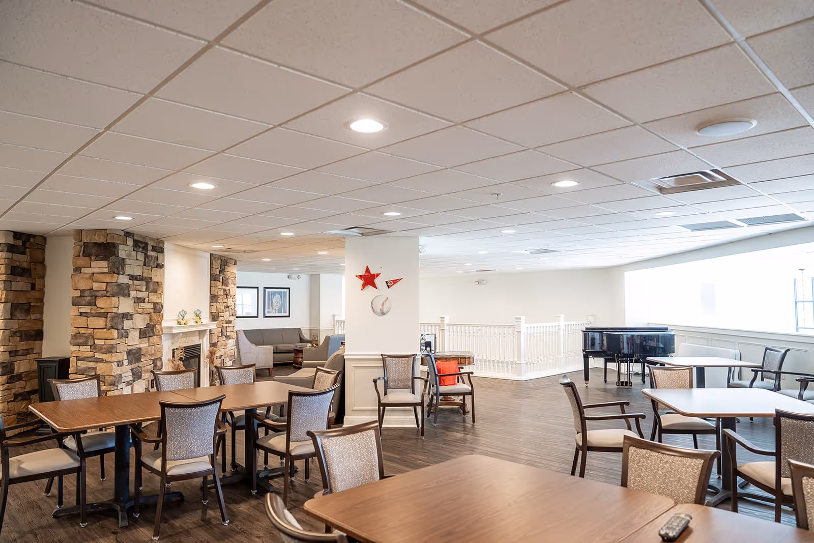 A spacious senior living common area with multiple wooden tables and chairs arranged for socializing or dining. The room features a stone fireplace on the left side, a black grand piano near a large window on the right, and a cozy seating area with sofas in the background. The ceiling has recessed lighting, and the floor is dark wood. A white railing separates part of the room, and decorative items including a red star and a baseball are mounted on a central column.