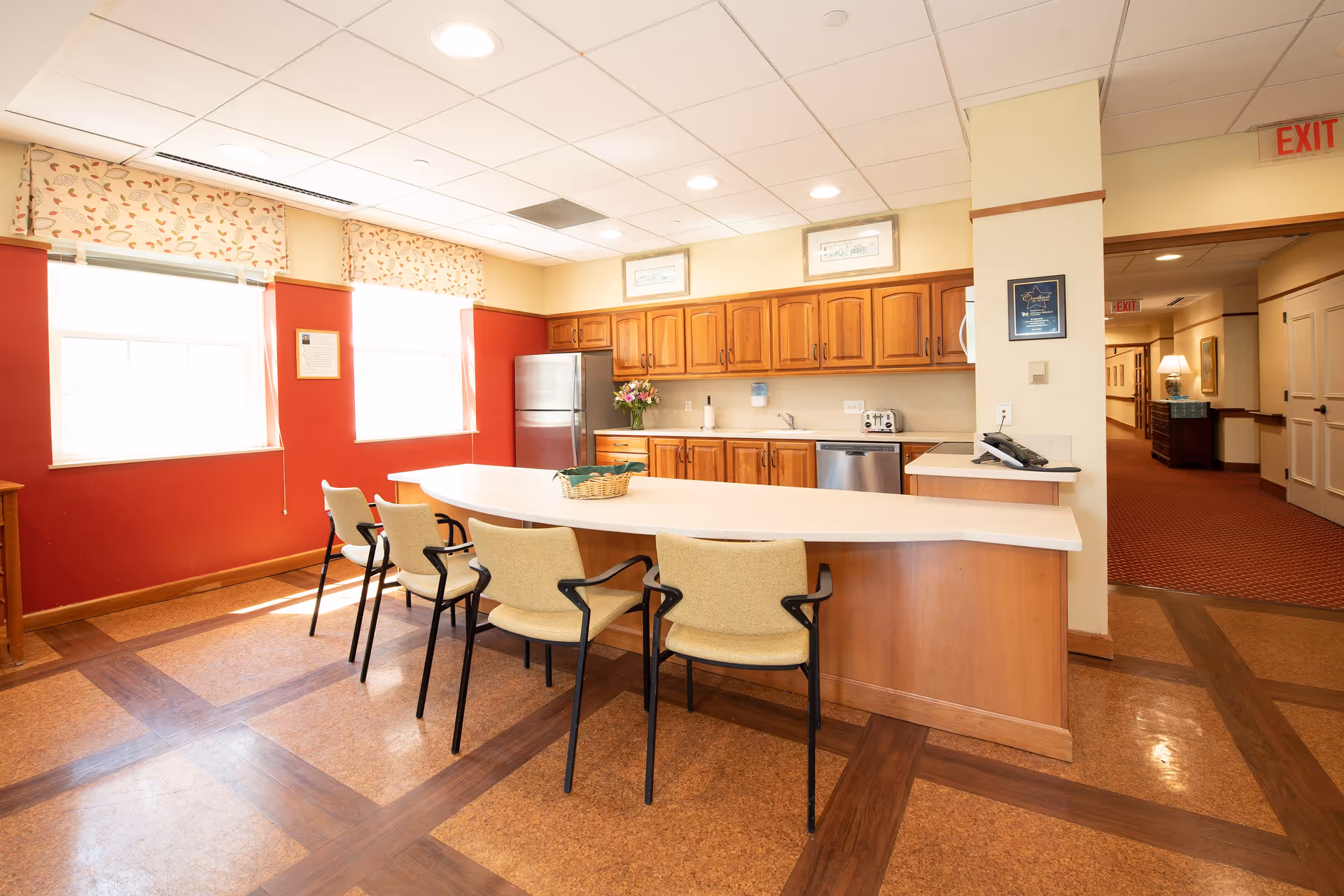 A bright kitchen area in a senior living facility with wooden cabinets, a stainless steel refrigerator, dishwasher, and toaster. There is a curved countertop with four beige chairs around it. Two large windows with floral valances let in natural light. A hallway with red carpet and exit signs is visible in the background.