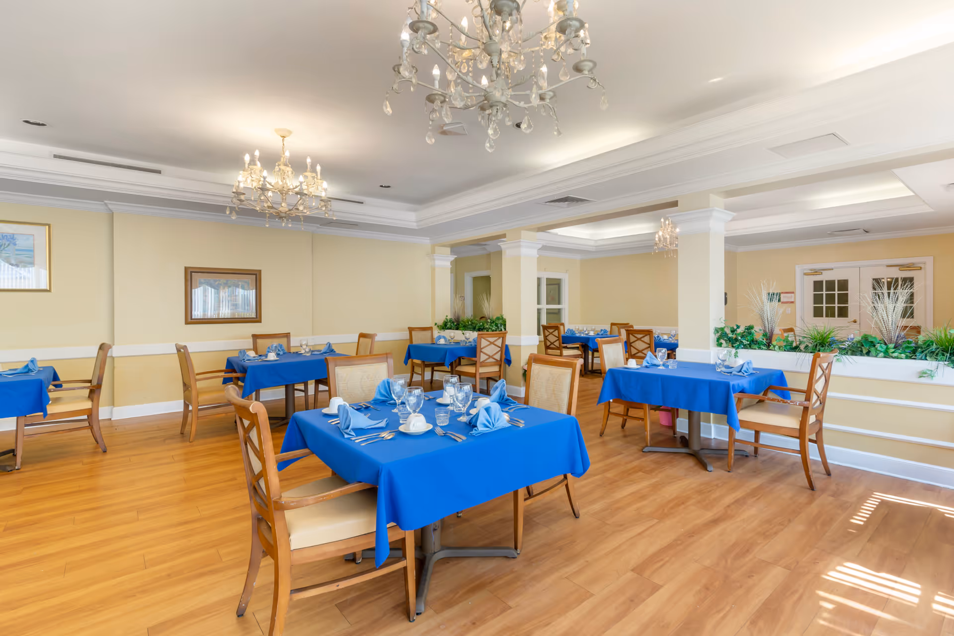 A bright dining room with wooden floors and cream-colored walls. Several tables are covered with blue tablecloths and set with glassware, napkins, and silverware. Wooden chairs with cushioned seats surround the tables. The room features chandeliers hanging from the ceiling and framed artwork on the walls. There are plants placed on a ledge dividing the room, and double doors are visible in the background.
