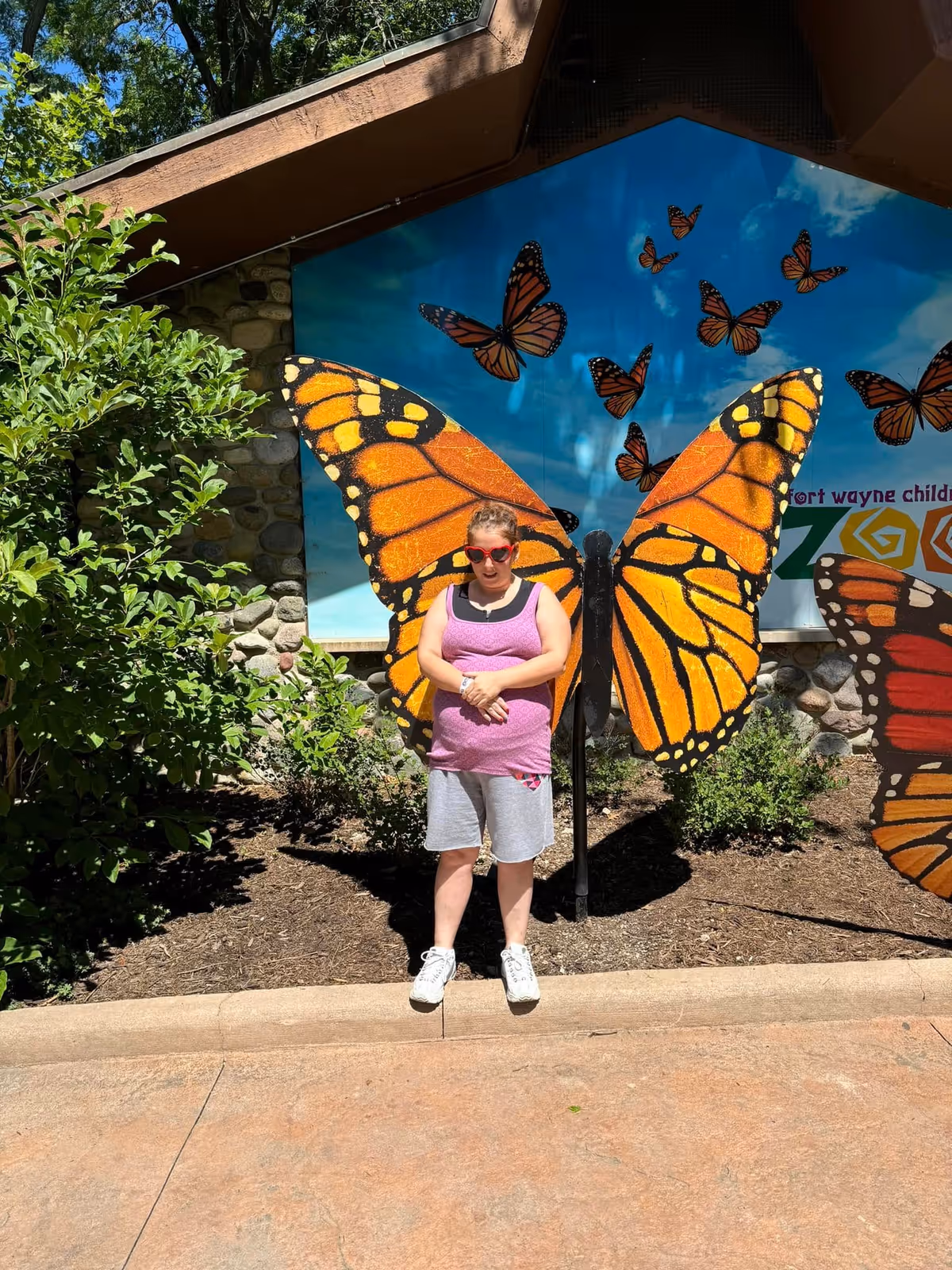 A person wearing a purple tank top, gray shorts, white sneakers, and red sunglasses stands in front of a large butterfly wing art installation outdoors, creating the illusion that the person has butterfly wings. The background includes a mural with multiple butterflies and some greenery on the left side.