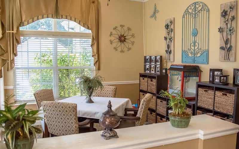 A cozy dining area with a table covered by a white tablecloth surrounded by four patterned chairs. There are green plants on the table and on a nearby ledge. The room has beige walls decorated with metal wall art and a window with beige curtains letting in natural light. There are storage units with woven baskets and a popcorn machine in the corner.