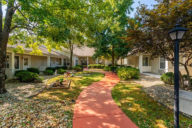 A landscaped outdoor courtyard with a red paved walkway winding through green grass, trees, and shrubs, surrounded by single-story beige buildings with white trim and windows. A black lamp post is visible on the right side of the walkway.