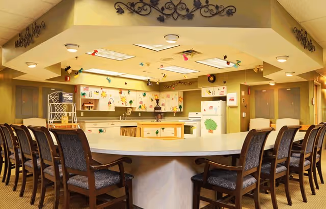 A large kitchen area with a curved counter surrounded by multiple cushioned chairs. The kitchen features white cabinets, a refrigerator decorated with colorful drawings, a stove, and a central island. The ceiling has recessed lighting and decorative hanging ornaments. The walls are adorned with various colorful children's artwork.