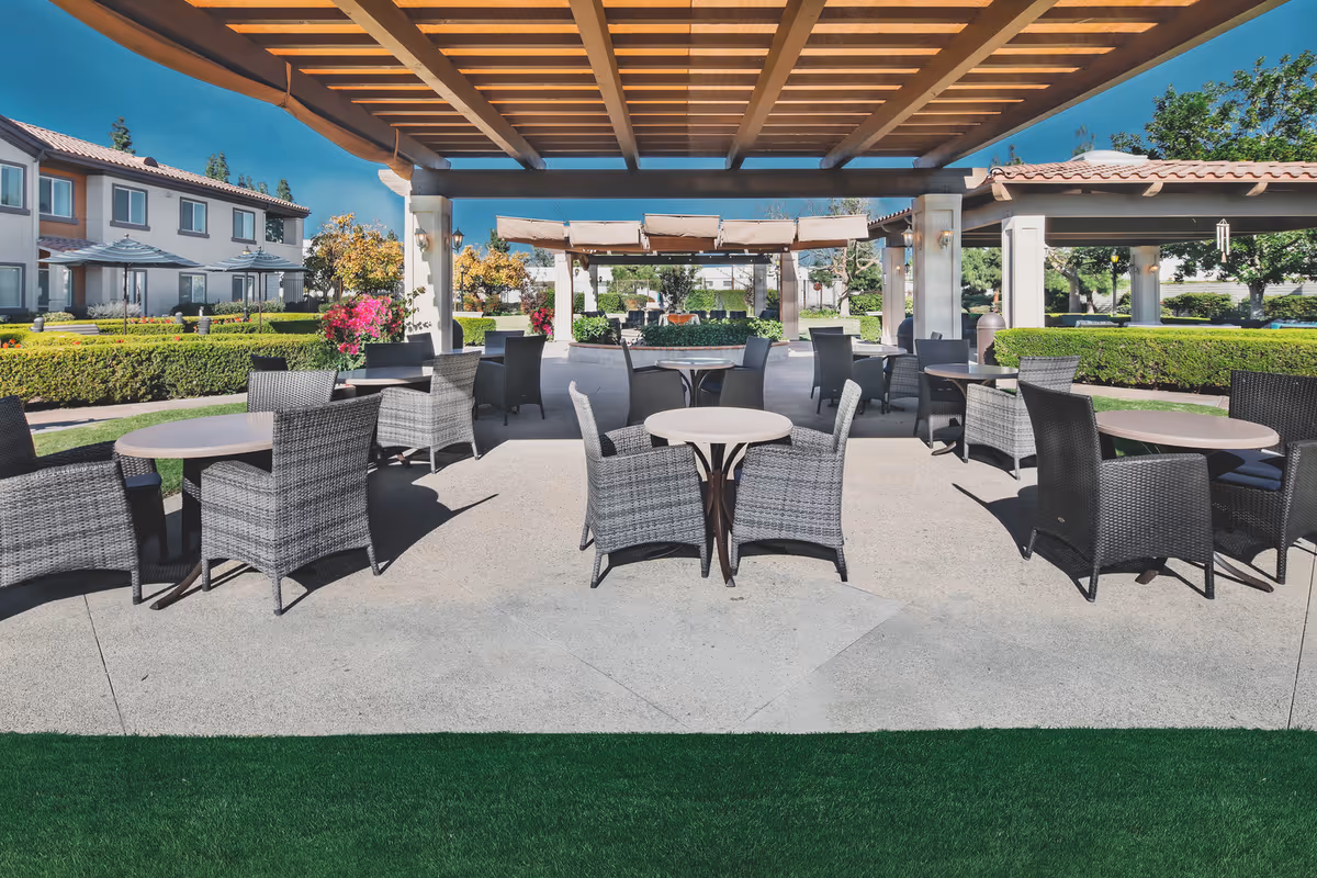 Covered outdoor patio with round tables and wicker chairs beneath a pergola in a landscaped courtyard.